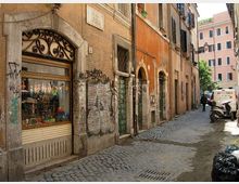 Vicolo stretto con pavimentazione in sampietrini, fiancheggiato da edifici storici intonacati con portoni ad arco, finestre con persiane e facciate segnate da graffiti. In primo piano si vede una vetrina con inferriata decorativa e, più avanti, un piccolo veicolo e sacchi della spazzatura lungo la strada.