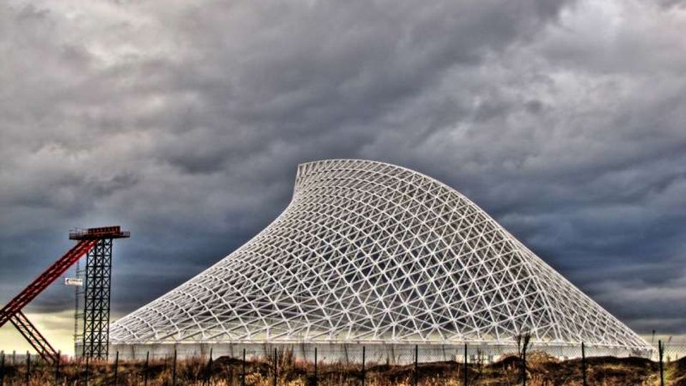Grande struttura architettonica bianca a rete reticolare, con profilo curvo simile a una collina artificiale, in un’area industriale recintata. In primo piano si vedono una strada con guardrail e vegetazione secca, mentre sullo sfondo il cielo è coperto da nuvole scure.