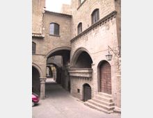 Cortile di un edificio storico in pietra, con portici ad archi e passaggi coperti che collegano le facciate. Si vedono finestre ad arco con inferriate, un grande portone in legno con scalini e una lanterna a muro su una parete in muratura.