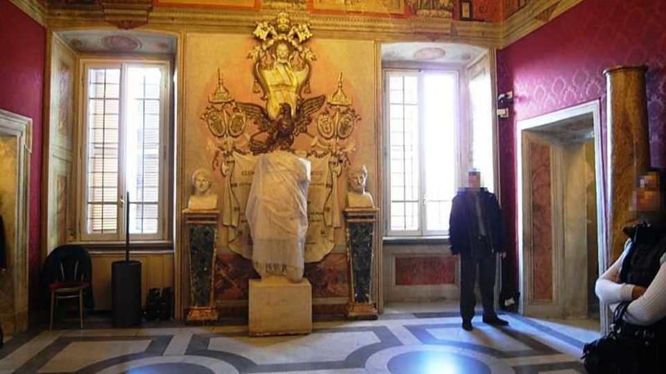 Interno di una sala storica decorata con affreschi e stucchi, con due grandi finestre e un lampadario a soffitto. Al centro della parete principale si trova una nicchia con scultura e busti laterali, mentre il pavimento presenta motivi geometrici in pietra.