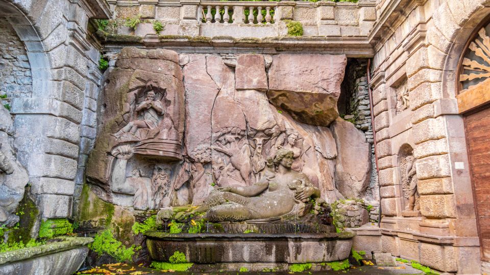 Cortile in pietra di un edificio storico con una grande fontana addossata al muro, decorata da bassorilievi scolpiti nella roccia e vasca rettangolare con muschio. Intorno si vedono pareti in blocchi di pietra, un arco e una balaustra superiore con vegetazione.