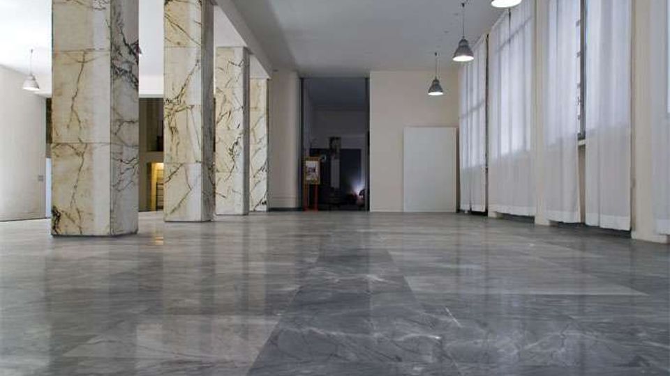 Ampia sala interna con pavimento in marmo grigio lucido e una fila di colonne rivestite in marmo venato. Sul lato destro ci sono grandi finestre con tende bianche e lampade a sospensione industriali lungo il soffitto.