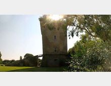 Torre in mattoni di forma quadrata in un’area verde, con un lungo muro in pietra che si estende lateralmente. Alberi e cespugli incorniciano la scena, con il sole che filtra tra le fronde sullo sfondo.