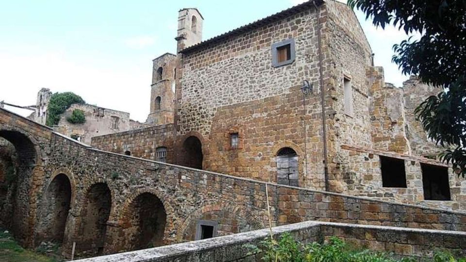 Edificio storico in pietra con facciate irregolari e piccole finestre, affiancato da un lungo muro con arcate in muratura e una rampa in salita. Sullo sfondo si vedono resti di strutture in pietra e vegetazione in un contesto rurale all’aperto.