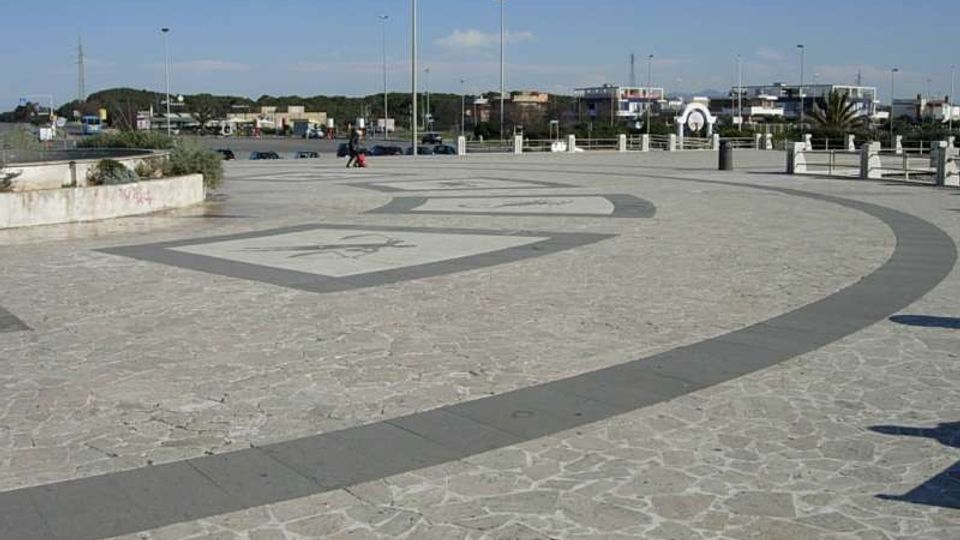 Ampia piazza pavimentata in pietra con motivi geometrici circolari e decorazioni incise, delimitata da lampioni e una balaustra. Sullo sfondo si vedono una strada con parcheggi e edifici bassi sotto un cielo sereno.