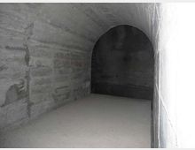 Interno di un tunnel sotterraneo in cemento grezzo, con pareti e soffitto curvi e pavimento spoglio. In fondo si vede un’apertura ad arco che conduce a una zona più buia.