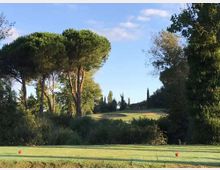 Campo da golf con ampio prato e area di tee in primo piano, circondato da vegetazione e alberi alti (inclusi pini a ombrello). Sullo sfondo si vede una leggera collina con cipressi e cielo sereno.