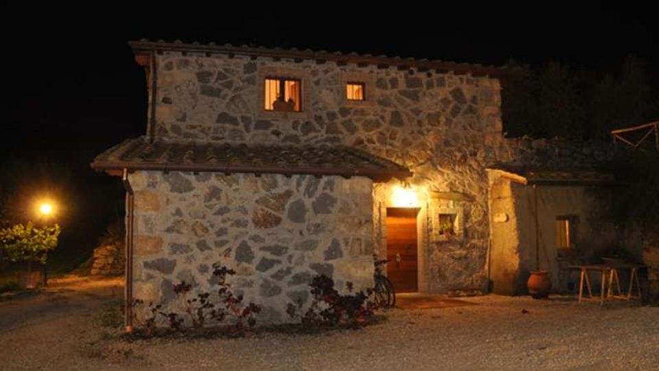 Casa rurale in pietra a due piani, vista di notte, con finestre illuminate e porta d’ingresso sotto una luce esterna. Davanti c’è un cortile in ghiaia con piante basse, un lampione sul lato sinistro e un piccolo annesso sulla destra.