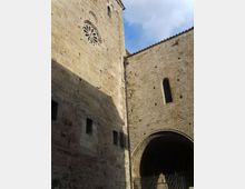 Facciata in pietra di un edificio storico, con rosone circolare decorato e piccole aperture rettangolari. A destra si vede un grande arco d’ingresso con cancello in ferro e una finestra stretta ad arco sotto un tetto in tegole, sullo sfondo cielo parzialmente nuvoloso.