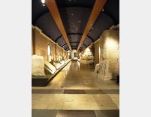 Interno di un museo archeologico con lunga galleria in pietra e mattoni, vetrine espositive illuminate lungo le pareti e grandi blocchi di rovine ai lati. Soffitto ad arco scuro con travi e luci incassate, pavimento in lastre di pietra con targhe e segnalazioni.