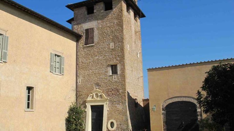 Cortile con prato davanti a un edificio storico in pietra con torre rettangolare e piccolo portale decorato. Ai lati si vedono corpi di fabbrica intonacati con finestre e un grande portone ad arco, sotto un cielo sereno.