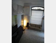 Interno di un soggiorno con pavimento in graniglia, divano a due posti e grande finestra ad arco con tenda a rullo. A sinistra ci sono un mobile TV basso, un tavolino con sedia e una lampada da terra accesa, con pareti bianche e un condizionatore vicino al soffitto.