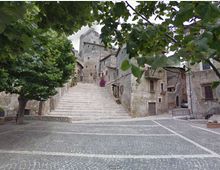 Scalinata in pietra che sale da una piazza lastricata verso un borgo medievale con edifici in muratura e una torre centrale. Alberi e rami in primo piano incorniciano la scena, con case addossate ai lati della scalinata.