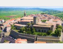 Veduta aerea di un borgo storico con edifici in pietra e tetti in coppi, dominato da un complesso fortificato con mura perimetrali, torre quadrata e chiesa con campanile. Sullo sfondo si estendono colline e campi coltivati.