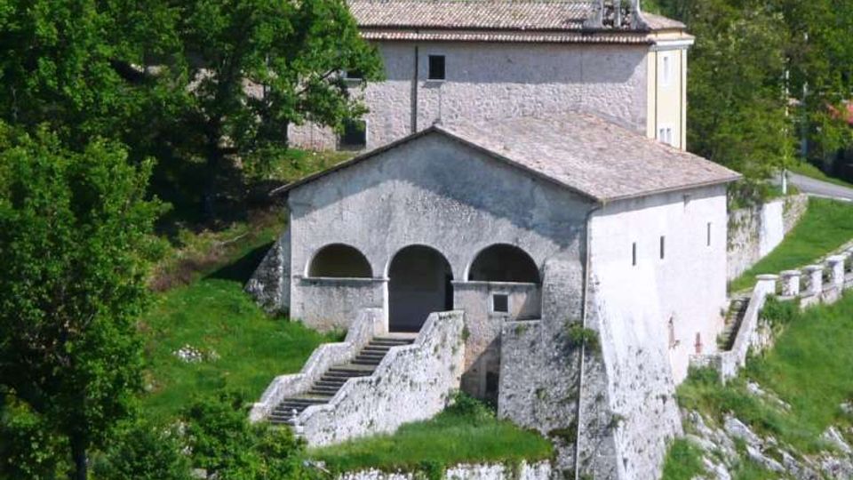 Chiesa rurale in pietra su un pendio erboso, con portico a tre arcate e scalinata che conduce all’ingresso. Sullo sfondo si vede un edificio annesso con campanile e tetto in coppi, circondato da alberi e muretti in pietra.
