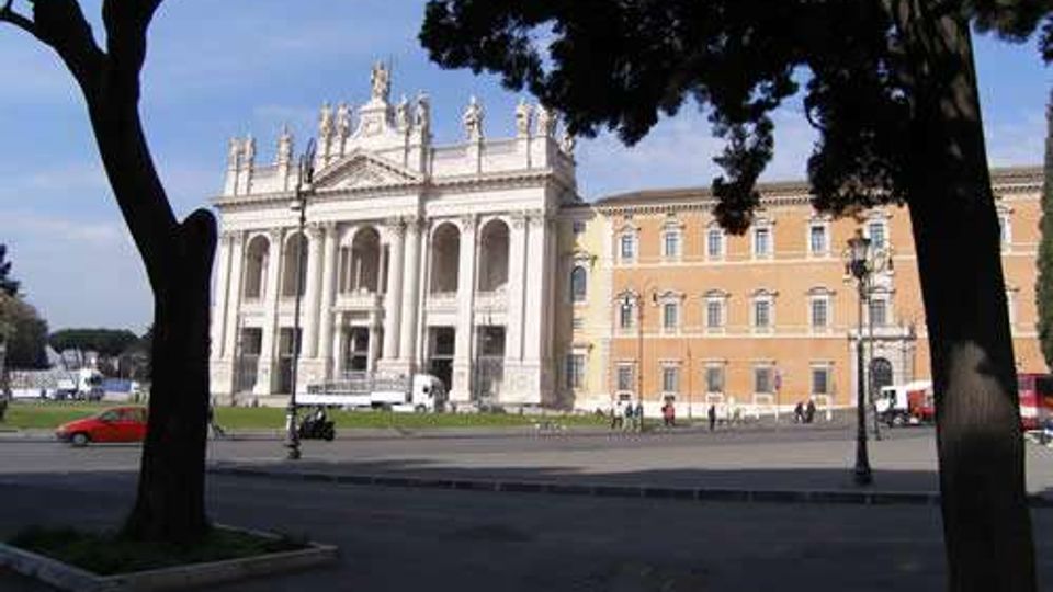 Facciata di un grande edificio storico in stile neoclassico con portico a colonne, frontone e statue sul tetto, affacciato su una piazza ampia. In primo piano si vedono alberi che incorniciano la scena e una strada con lampioni e alcune auto parcheggiate.