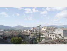Veduta panoramica di un borgo su una collina con case dai tetti in coppi e edifici in pietra, tra cui un campanile e grandi strutture storiche. Sullo sfondo si estendono colline e montagne sotto un cielo parzialmente nuvoloso.