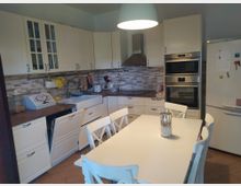 Cucina domestica luminosa con mobili bianchi a parete, piano di lavoro in legno e paraschizzi a mosaico in tonalità beige e grigie. In primo piano c’è un tavolo da pranzo bianco con sedie, mentre sul lato destro si vedono forno e microonde incassati e un grande frigorifero.