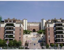 Complesso residenziale moderno con due palazzi in mattoni e cemento ai lati, dotati di balconi, e un edificio centrale più alto sullo sfondo. In primo piano una scalinata ampia con murales sui muri di contenimento conduce a un arco d’ingresso, con alberi e strada alla base.