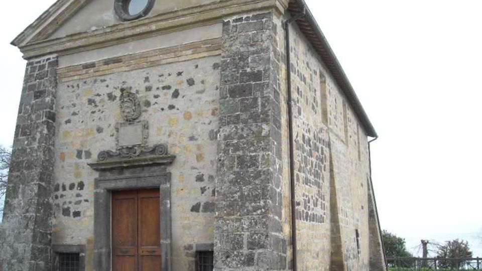 Facciata di una piccola chiesa in pietra e intonaco, con frontone triangolare e finestra circolare in alto, portale centrale in pietra e porta in legno. L’edificio è isolato su un’area erbosa, con muri laterali in muratura a vista e pochi elementi decorativi.
