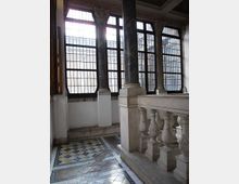 Interno di un edificio storico con ampie finestre a riquadri in ferro, colonne in pietra con capitelli decorati e pavimento a mosaico geometrico. In primo piano si vede una balaustra in marmo lungo una scala o un ballatoio.