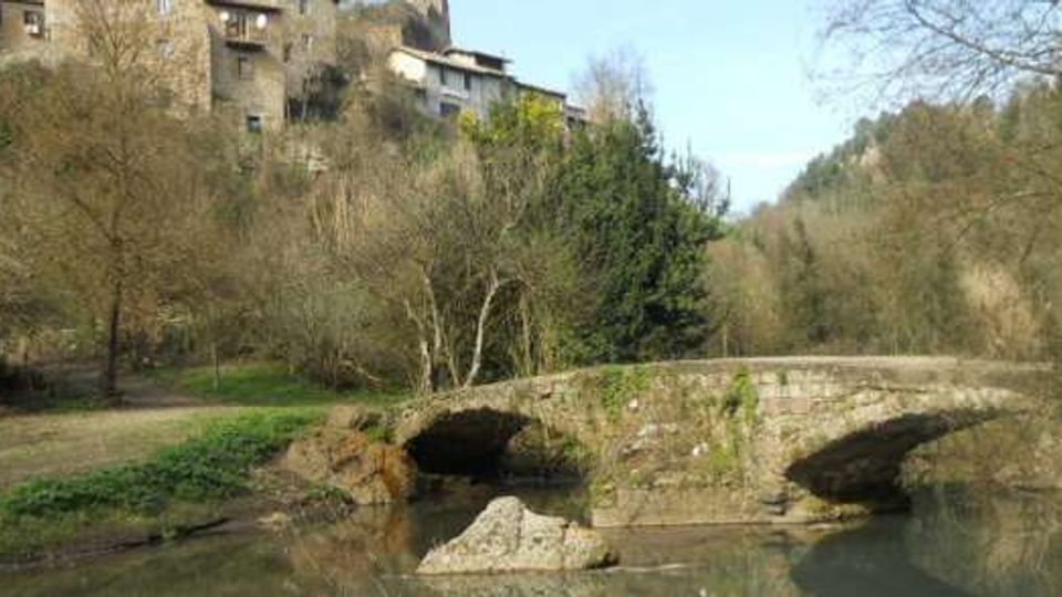 Ponte in pietra a due arcate che attraversa un fiume calmo, circondato da alberi spogli e vegetazione lungo le rive. Sullo sfondo si vedono case in pietra su un pendio e colline boscose.