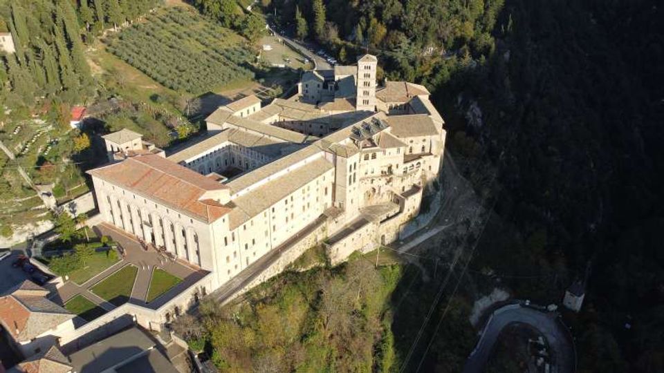 Veduta aerea di un grande complesso monastico in pietra chiara con edifici a tetto in coppi e una torre campanaria, disposto attorno a cortili interni. La struttura è costruita sul bordo di un pendio boscoso, con oliveti e strade di accesso nelle vicinanze.