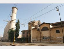 Vista stradale urbana con un grande edificio industriale in muratura e un’alta ciminiera cilindrica bianca, affiancati da un fabbricato basso con tetto in tegole e pareti con graffiti. In primo piano si vedono binari del tram, cavi aerei, pali e un lampione lungo la strada.