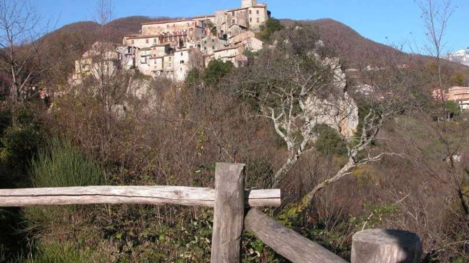 Borgo storico arroccato su una collina, con case in pietra addossate tra loro e una torre in cima. In primo piano si vede una staccionata in legno e vegetazione invernale, con colline boscose sullo sfondo sotto un cielo sereno.