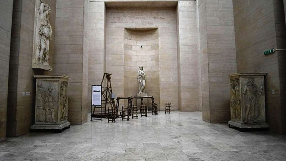 Interno di una grande sala museale in pietra chiara, con pavimento in lastre e alto lucernario a soffitto. Al centro una statua su piedistallo e attrezzature sceniche in legno, affiancate da due grandi basamenti scolpiti e una nicchia sul fondo.