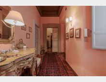 Corridoio interno di una casa con pareti rosa e soffitto a cassettoni scuro, pavimento in parquet e lungo tappeto persiano centrale. A sinistra c’è una consolle decorata con grande specchio e lampada, mentre a destra si vedono applique, quadretti incorniciati e un citofono vicino a una finestra; in fondo una porta conduce ad altre stanze.