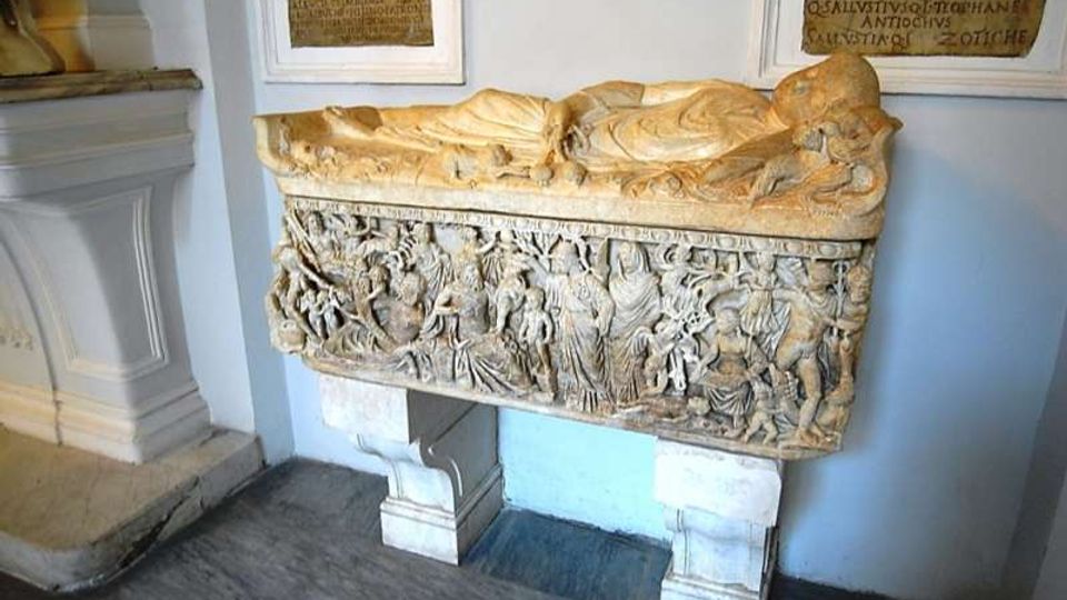 Sarcofago in pietra esposto in una sala museale, con coperchio scolpito a figura distesa e pannello frontale riccamente decorato a bassorilievo con scene e personaggi. Sul muro chiaro dietro sono montate varie lapidi con iscrizioni.