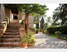 Cortile esterno in pietra con scala di accesso a un edificio rustico intonacato, affiancata da vasi in terracotta con piante. Viale pavimentato che prosegue tra alberi e siepi, con panchina e ingresso con cancello sullo sfondo.