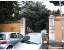 Ingresso con cancello in ferro battuto tra pilastri in pietra e muro intonacato color ocra, affacciato su una strada con auto parcheggiate; dietro il cancello si vede un’area verde con alberi alti e fitti.