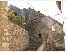 Scalinata in pietra in un vicolo stretto di un borgo antico, affiancata da alti muri e case in muratura grezza. Si vedono una ringhiera in ferro battuto, un lampione a parete e cavi aerei tra gli edifici.