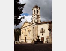 Chiesa in stile barocco con facciata color ocra e campanile in pietra sormontato da cupola, affacciata su una piccola piazza con una fontana centrale. Sullo sfondo si vedono edifici adiacenti e alberi, sotto un cielo nuvoloso.