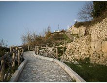 Sentiero in pietra in salita con parapetti in legno, che serpeggia tra muretti a secco e terrazzamenti erbosi su un pendio. In alto si intravedono lampioni e costruzioni lungo il percorso, con vegetazione rada ai lati.
