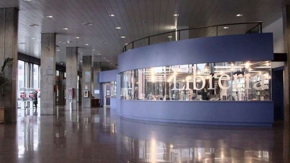 Ampio atrio interno moderno con pavimento lucido, colonne in cemento e soffitto a pannelli con luci incassate. Al centro c’è un volume curvo blu con grandi vetrate e la scritta “caffetteria”, vicino a finestre a tutta altezza lungo il lato sinistro.