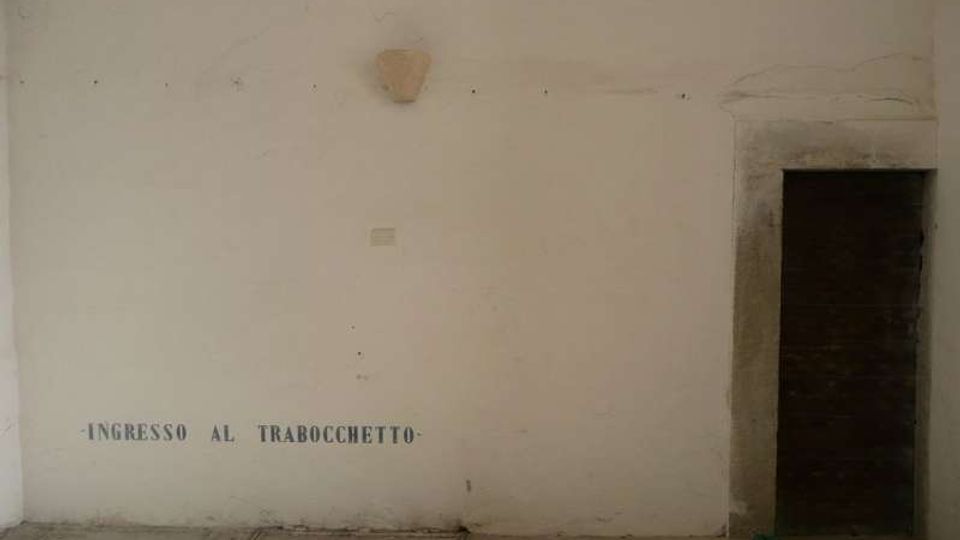 Parete interna intonacata con scritta “INGRESSO AL TRABOCCHETTO” vicino al pavimento e una piccola applique al centro. A destra si vede un’apertura con cornice in pietra che conduce a un vano buio, con pavimento in lastre e un pannello metallico a filo.