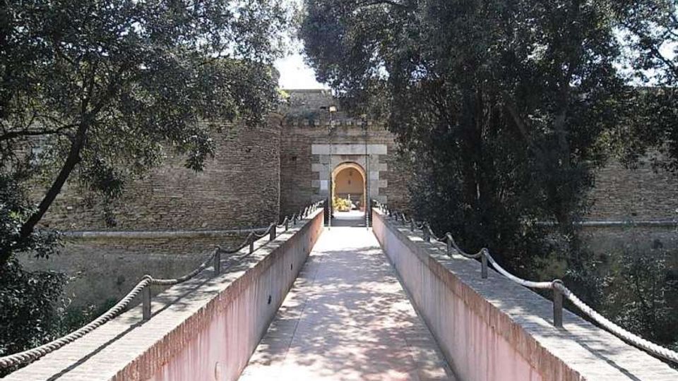 Passerella pedonale in muratura con corrimano in corda che conduce a un arco d’ingresso in pietra su un alto muro fortificato. Ai lati si vedono alberi e vegetazione che incorniciano il percorso.