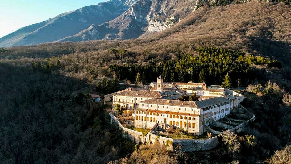Veduta aerea di un grande complesso monastico fortificato con edifici in pietra e intonaco chiaro, tetti in tegole e un piccolo campanile, racchiuso da mura. Il complesso sorge in una valle boscosa ai piedi di montagne rocciose.