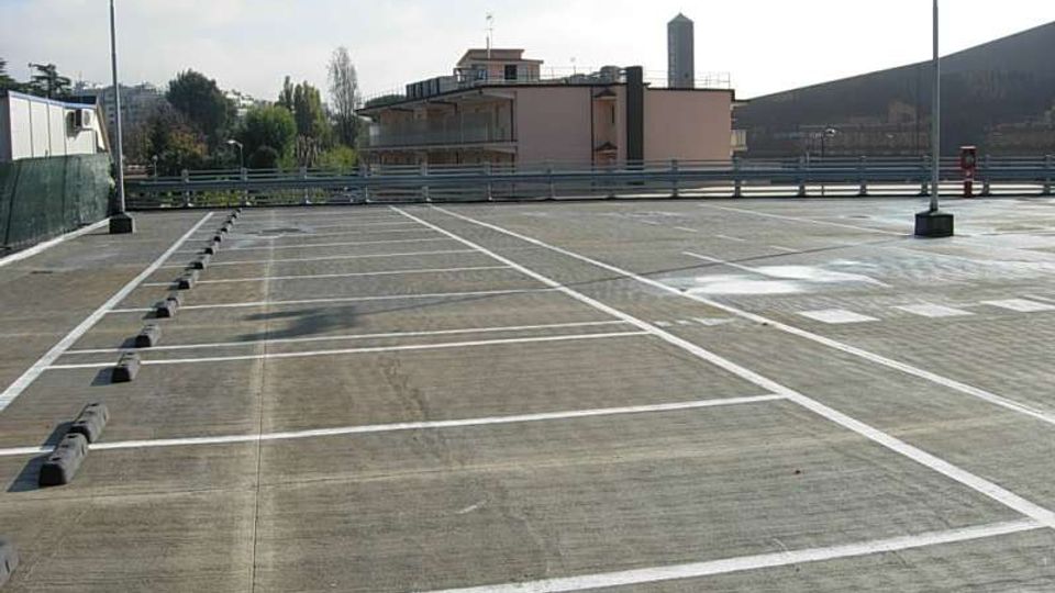 Parcheggio all’aperto su un piano rialzato, con ampie strisce bianche di delimitazione dei posti e cordoli di gomma. Sullo sfondo si vedono una ringhiera, edifici bassi, alberi e alcuni lampioni sotto un cielo chiaro.