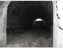 Interno di un tunnel o passaggio sotterraneo in cemento con volta a botte, pareti scure e pavimento coperto di macerie e detriti. In fondo si vede un’apertura ad arco che lascia entrare luce dall’esterno.