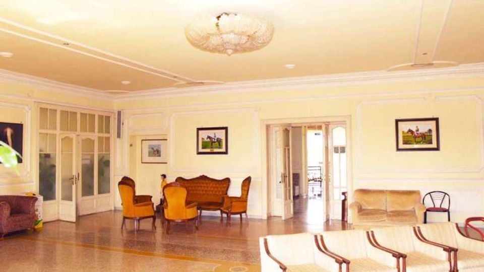 Ampia sala interna in stile classico con pareti chiare decorate da cornici, lampadario centrale e pavimento in graniglia con motivi. Sono presenti divani e poltrone disposte come area lounge, quadri alle pareti e grandi porte vetrate che collegano ad altre stanze.