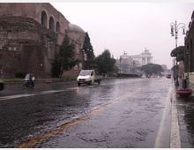 Strada urbana bagnata dalla pioggia con pavimentazione in sampietrini e lampioni lungo il marciapiede. A sinistra si vedono grandi mura in mattoni con arcate e una cupola, mentre sullo sfondo appare un edificio monumentale su una collina.
