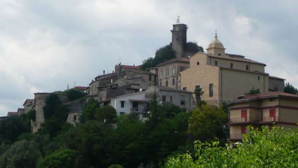 Veduta di un borgo collinare con case in pietra e intonaco addossate al pendio, circondate da vegetazione. In cima spiccano una torre cilindrica medievale e una chiesa con campanile sotto un cielo nuvoloso.