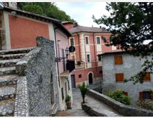 Vicolo in un borgo collinare con una scalinata in pietra e un muretto che conduce a case ravvicinate. Gli edifici includono una casa intonacata rosa con balconcino e un edificio in pietra con persiane in legno, circondati da alberi e vegetazione.