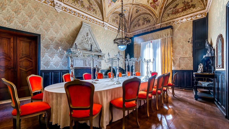 Sala da pranzo storica con grande tavolo ovale apparecchiato e sedie imbottite rosse, pavimento in legno e boiserie scura alle pareti. Soffitto a volte affrescato, grande camino in pietra scolpita, lampadario centrale e ampia finestra con tende che illumina l’ambiente.