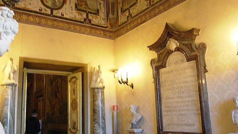 Interno di una sala storica con soffitto a cassettoni decorato e fregi dipinti lungo il perimetro, illuminata da lampadari e applique. Sulle pareti si vedono busti e monumenti marmorei, una grande lapide commemorativa e un portale con ante decorate che conduce a un ambiente adiacente.