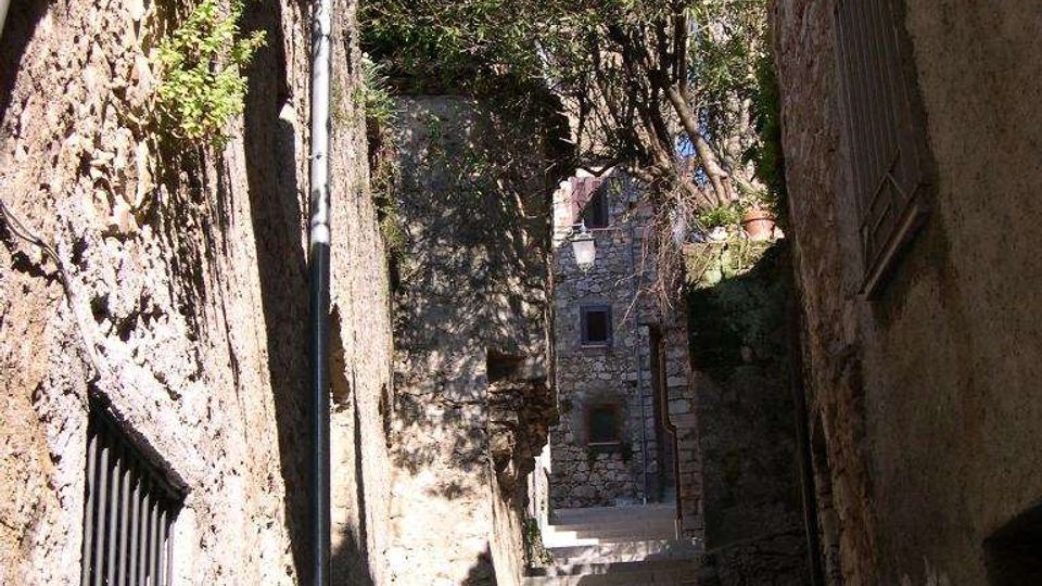 Scalinata in pietra in un vicolo stretto tra edifici in muratura, con pareti ruvide e finestre con grate. Un lampione in ferro sporge sopra le scale, con piante rampicanti e alberi che incorniciano la parte alta del passaggio.